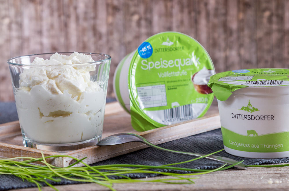 Ein Glas frischer Quark und zwei verschlossene Quarkbecher mit Dittersdorfer Logo, dekorativ auf einem Holzbrettchen angerichtet