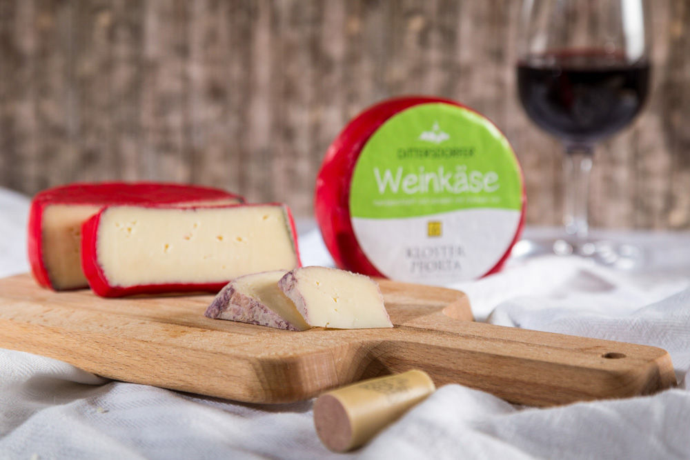 Ein angeschnittener Laib Weinkäse liegt auf einem Holzbrett, daneben steht ein Glas Rotwein