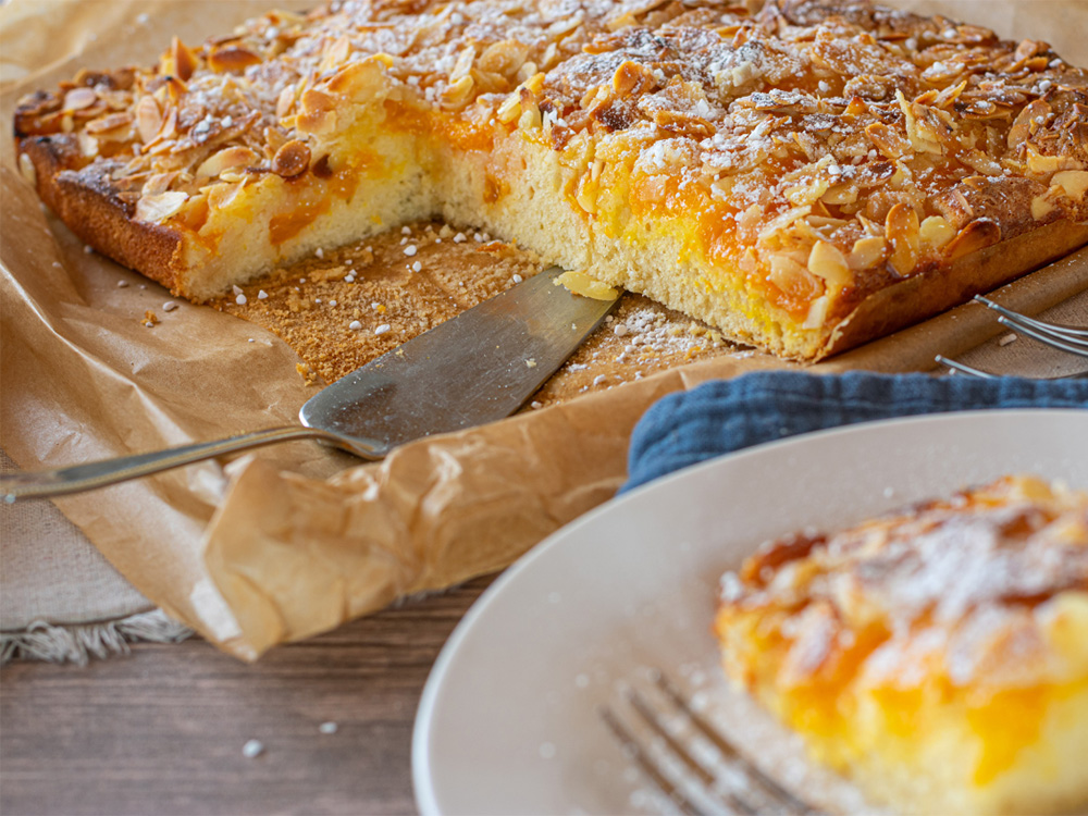Ein angeschnittener Blechkuchen mit Mandeln sowie ein Stück Kuchen auf einem Teller mit einer Gabel