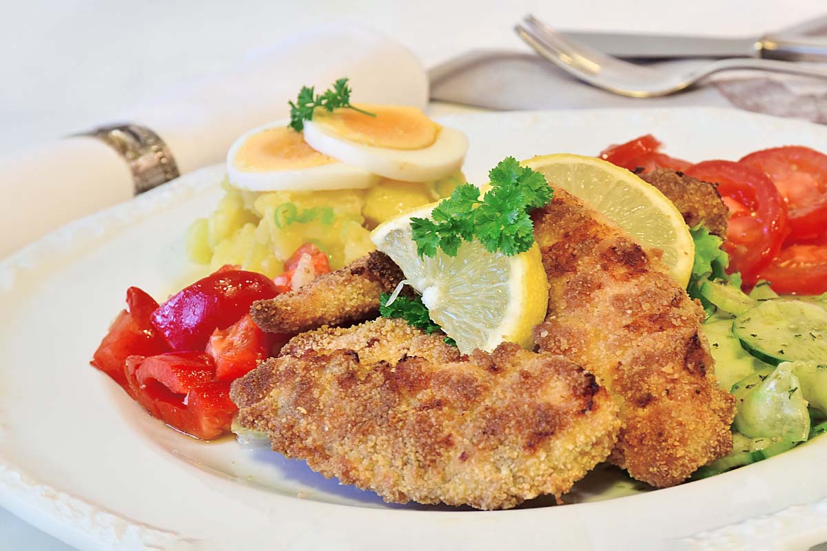 Ein Bild eines Mittagessens. Angerichtet wurden ein Schnitzel mit Zitronenscheibe, Kartoffelsalat mit Ei und verschiedene Beilagen