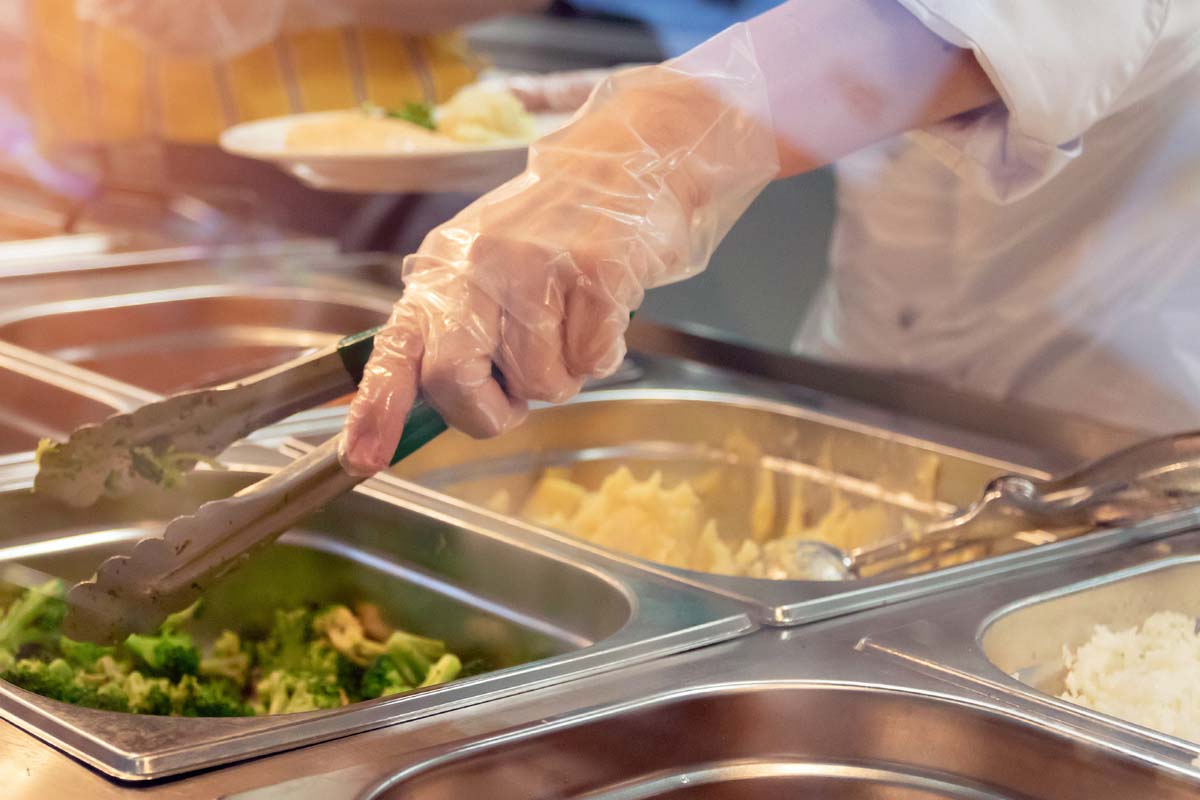 Das Bild zeigt die Hand einer Servicekraft bei der Essensausgabe. Es sind verschiedene Wärmebehälter mit Speisen zu sehen