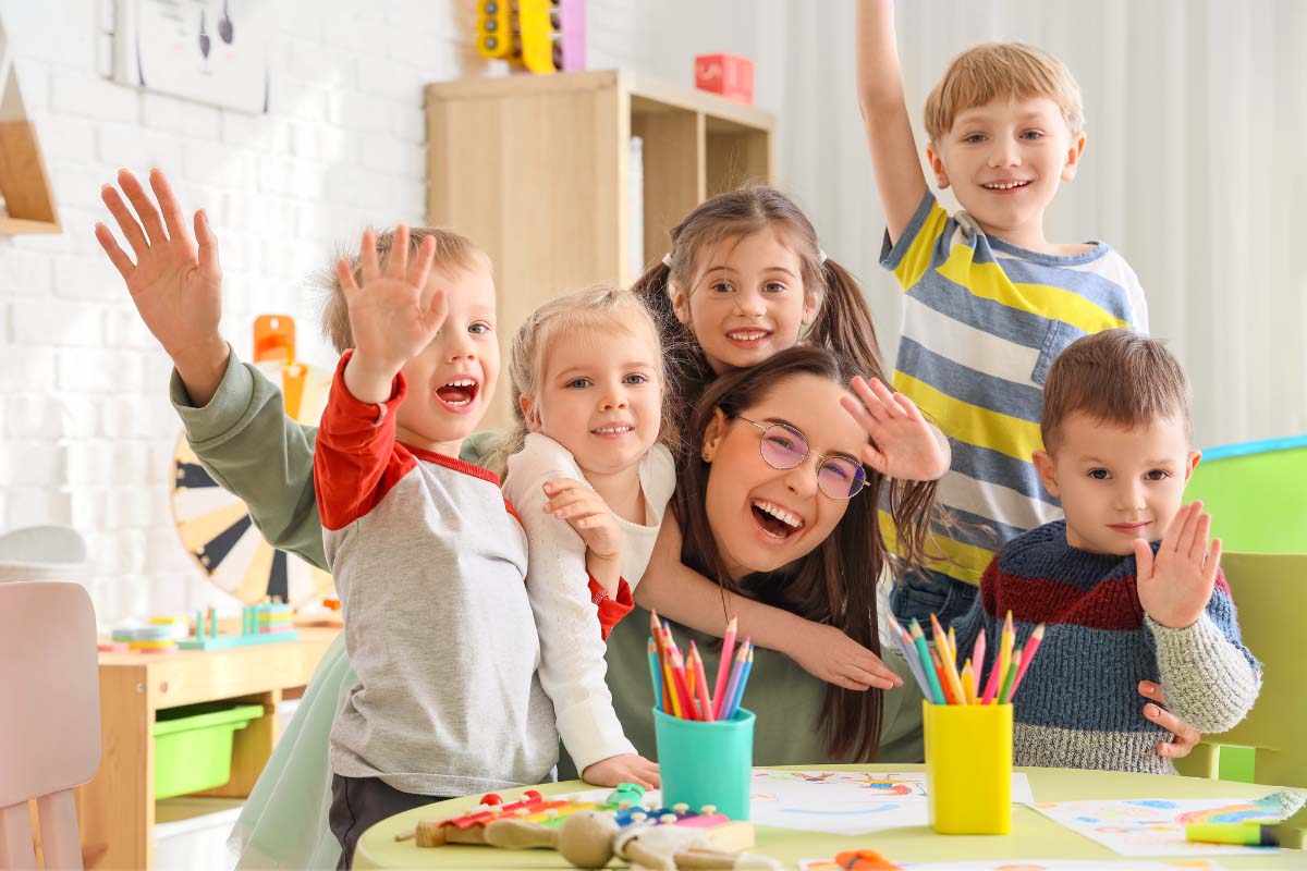 Das Bild zeigt eine Erzieherin eines Kindergartens mit 5 Kindern, die in die Kamera winken. Auf dem Tisch stehen Behälter für Buntstifte und Malpapier