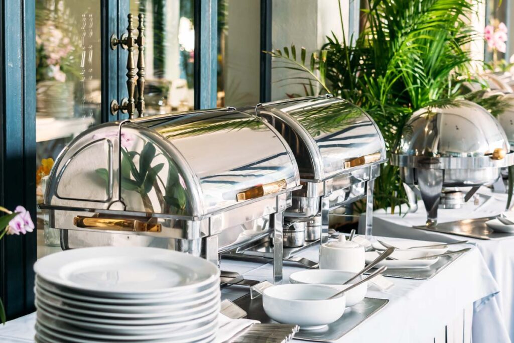Ein Buffet mit Speisenwärmern aus Edelstahl und weißem Geschirr Im Hintergrund steht eine größere Zimmerpflanze