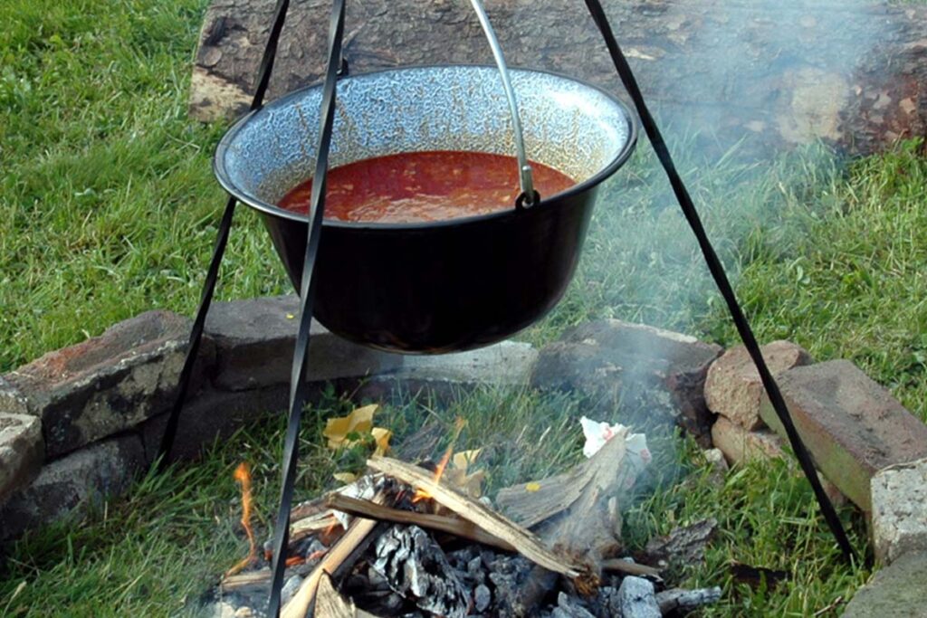 Über einem Lagerfeuer hängt, in einem dreibeinigen Gestell, ein Topf mit Suppe